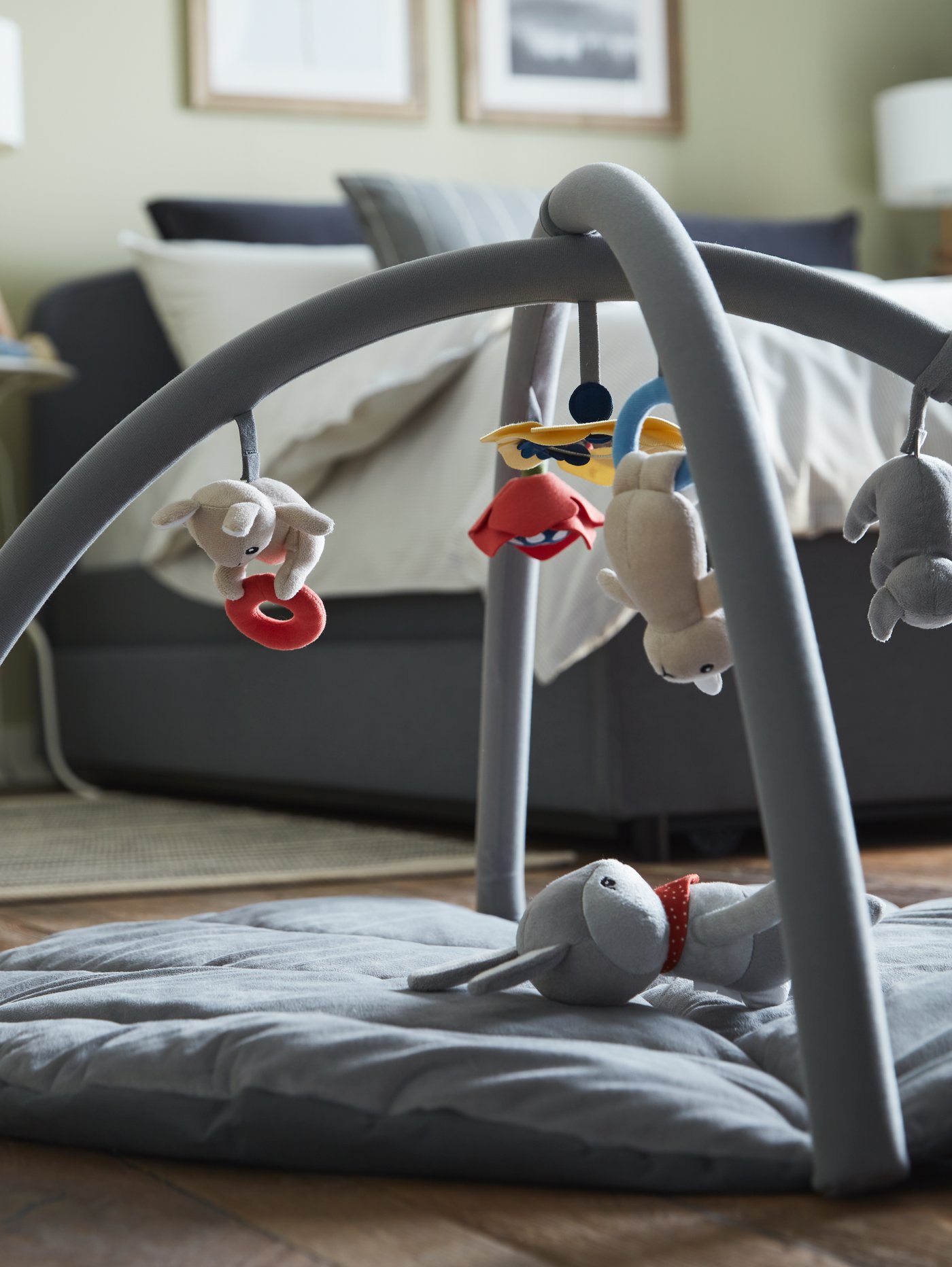 A GULLIGAST soft toy shaped like a rabbit lies in a multicolour GULLIGAST baby gym near a grey HAUGA upholstered bed.