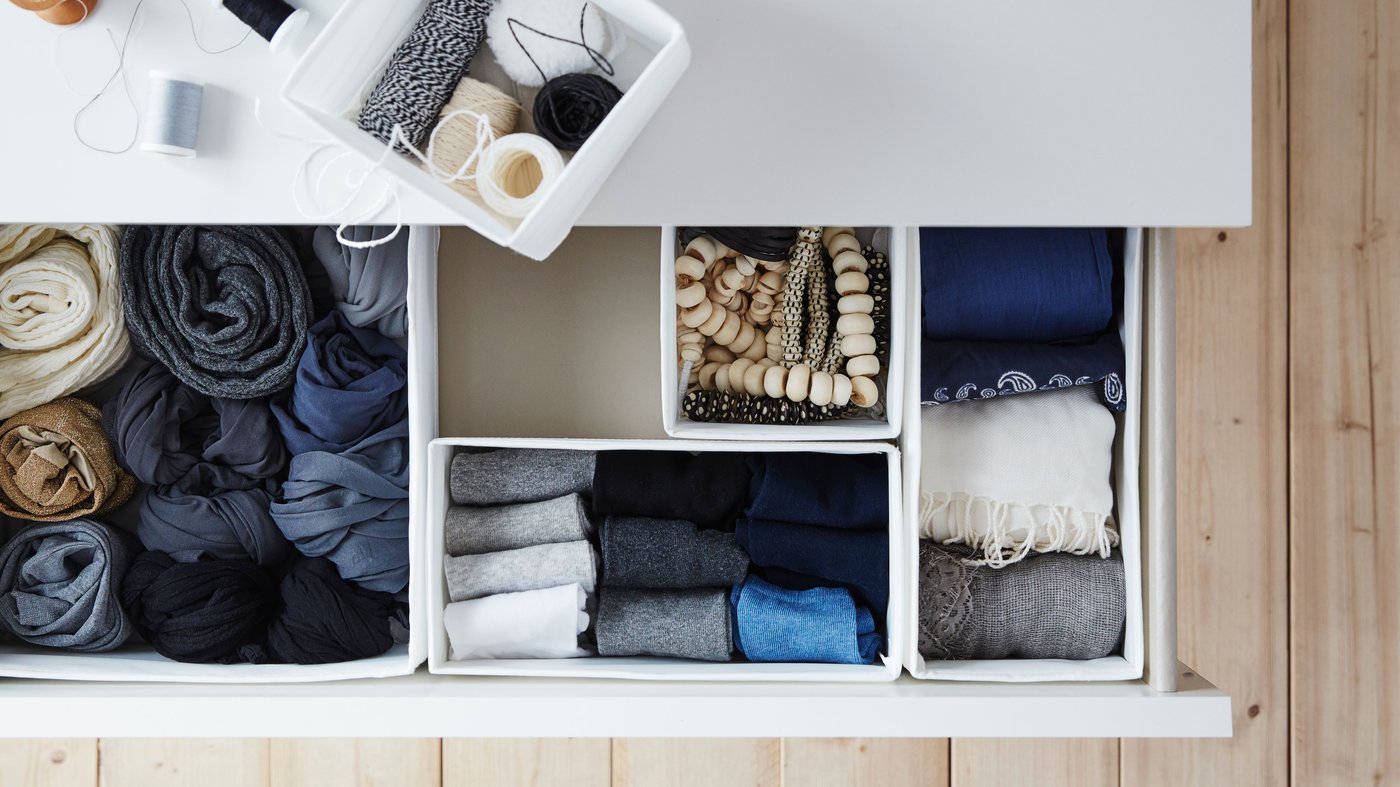 Es sind fünf weiße SKUBB Boxen mit Kleidung und Accessoires zu sehen. Vier davon befinden sich in einer offenen Schublade einer weißen MALM Kommode. Eine Box liegt oben auf der Kommode.