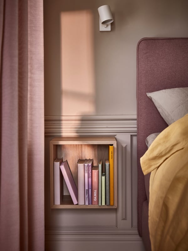 Ein würfelförmiges Regalelement mit Büchern darin hängt neben einem rosefarbenen Boxspringbett an einer mit Holz-Kassetten verzierten Wand im Stil der Jahrhundertwende.
