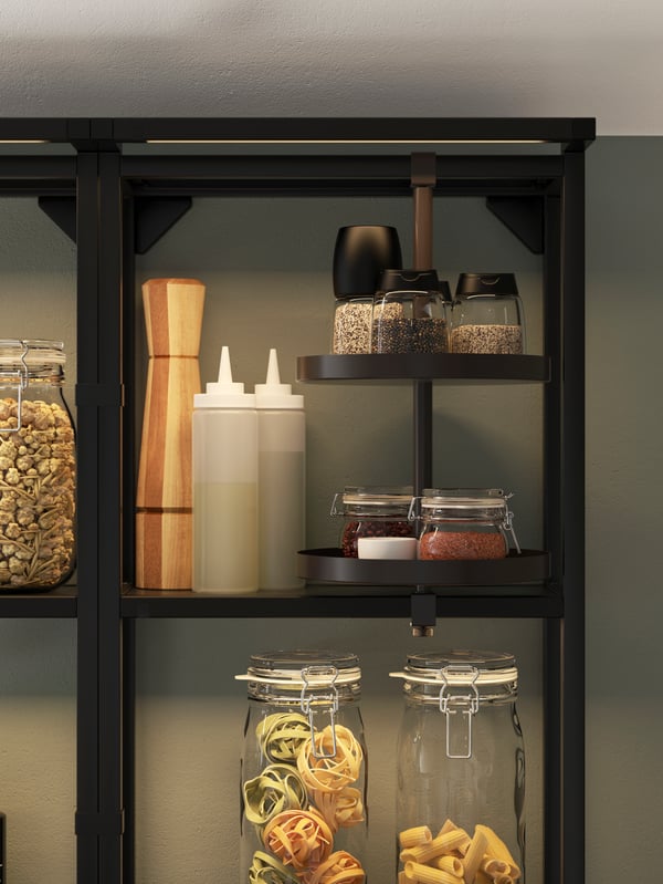 A black open shelf with integrated lighting and a swivel shelf, storing glass jars with pasta, spice mills, and condiments.