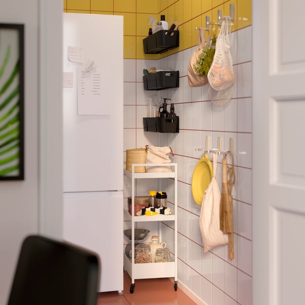 A tight corner of a kitchen with a NISSAFORS trolley in white and KUNGSFORS net bags in cotton holding diverse items.