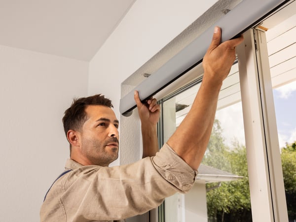 A man in a beige shirt is installing a grey window blind or curtain rod above a glass door or large window.