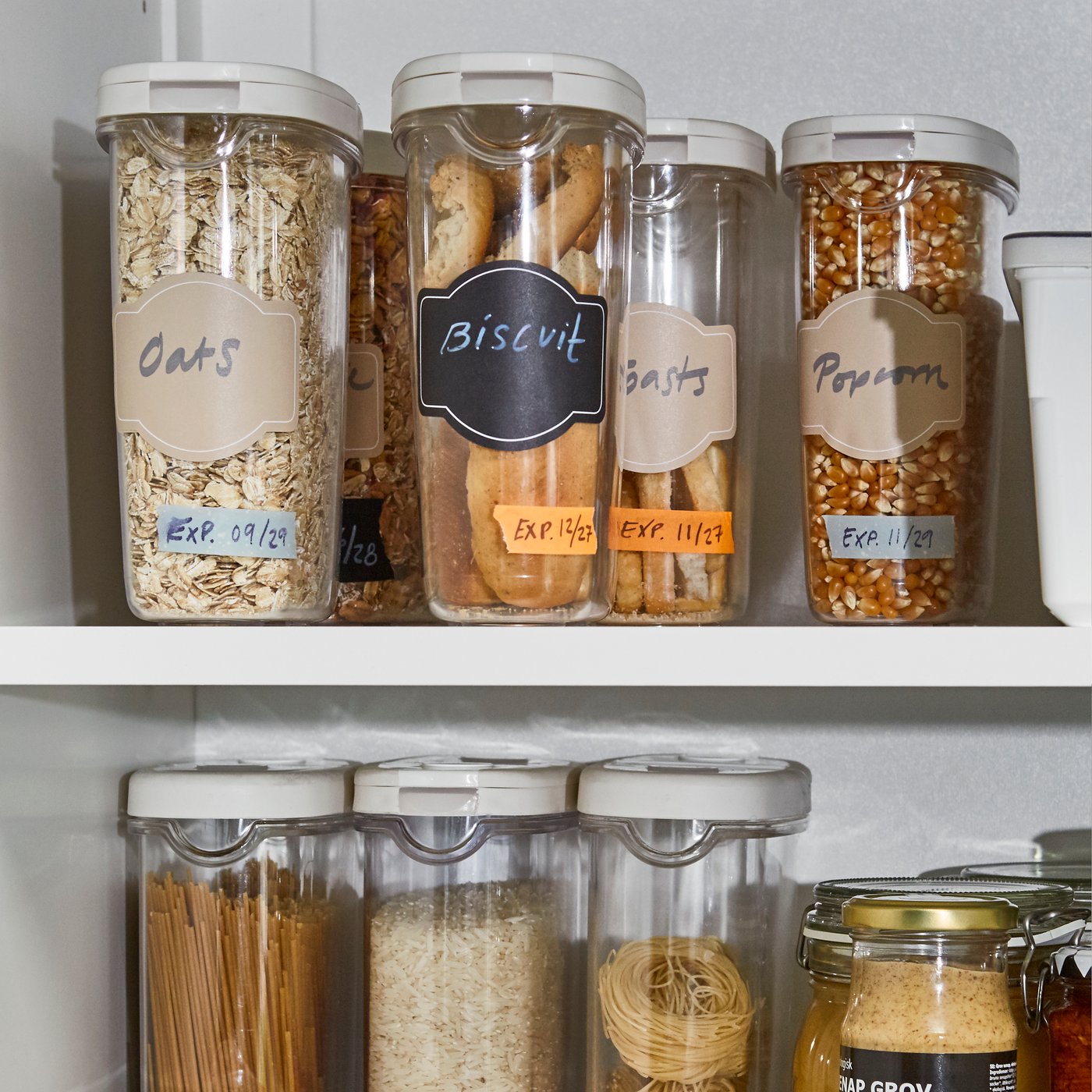 Des boîtes de conservation en plastique de différentes tailles munies d’étiquettes sur deux tablettes dans une armoire de cuisine.