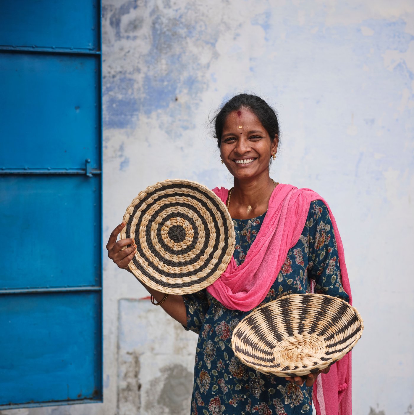 A person in a pink shawl stands before a weathered white wall. They are smiling and holding up the MÄVINN wall decoration. 