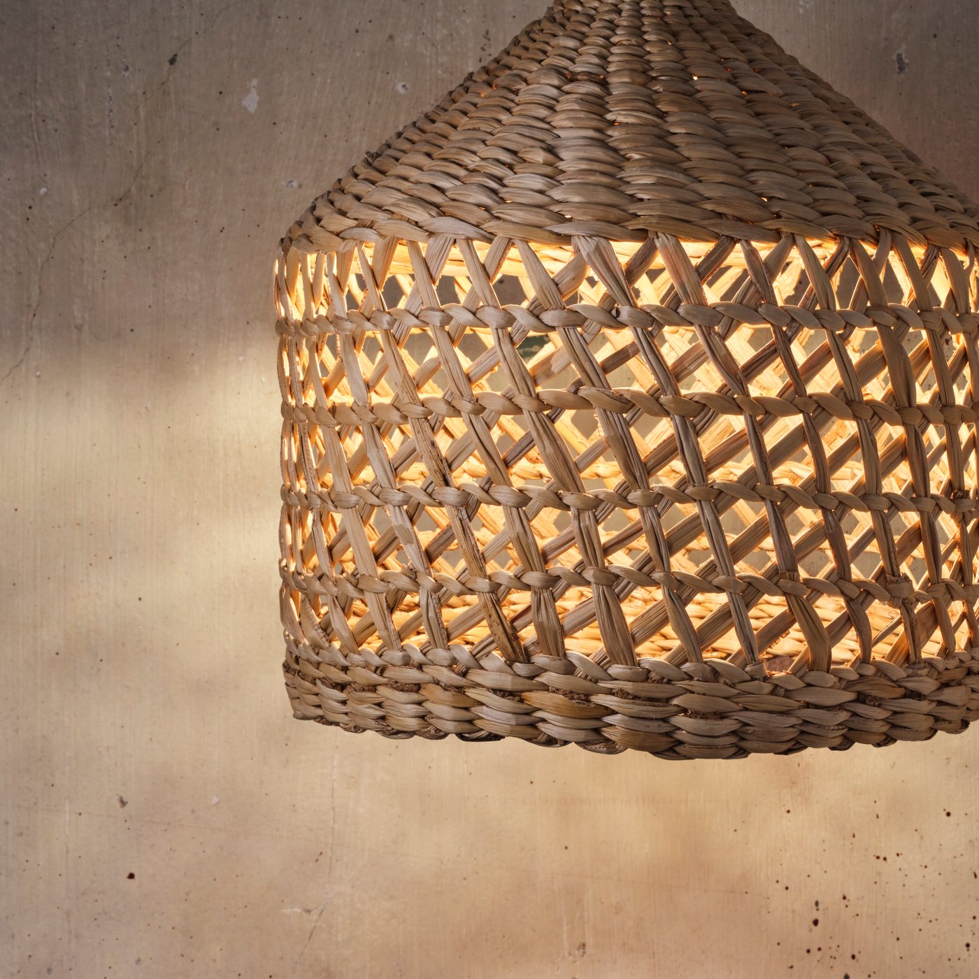 MÄVINN lampskärm för taklampa hänger framför en grå betongvägg. Lampan är tänd, vilket framhäver det utskurna mönstret.