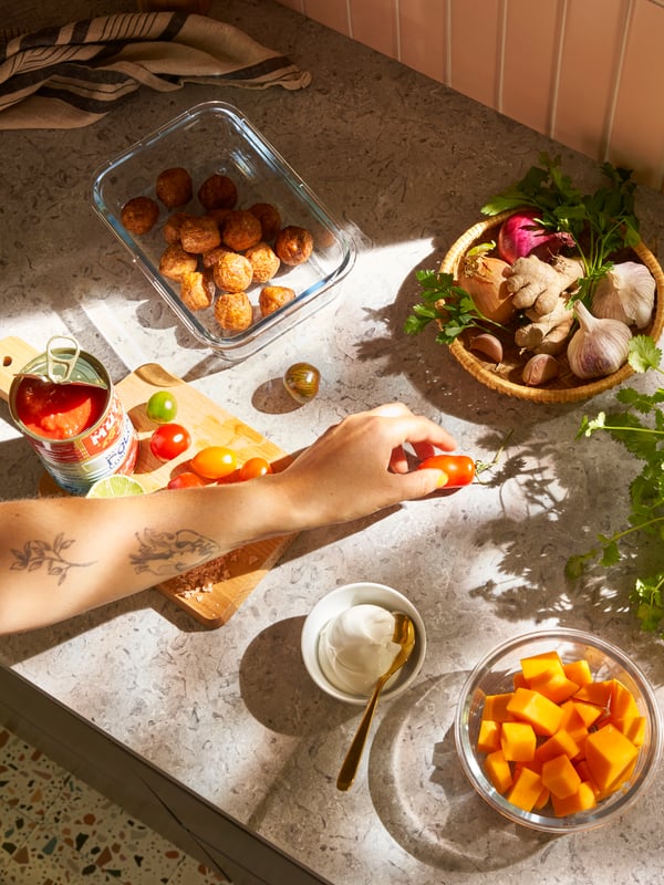 Una mano toma un tomate en una barra de cocina con un recipiente de cristal con albóndigas de proteína vegetal HUVUDROLL y otros ingredientes.