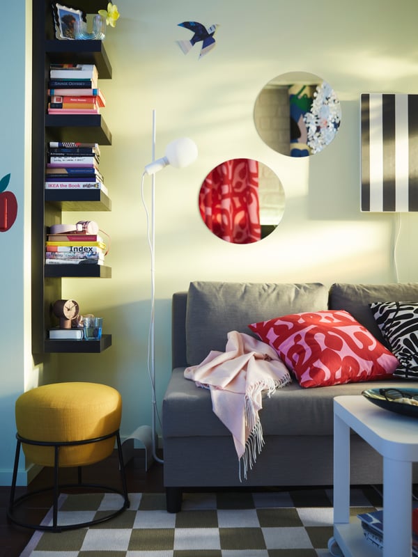 A colourful corner with a black-brown LACK wall shelf with book stacks, an ÄLVDALEN sofa-bed and a yellow EMMISLÖV pouffe.