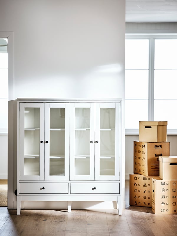 A room with white walls and floorboards that is empty except for an IDANÄS cabinet with glass bi-fold doors and some boxes.