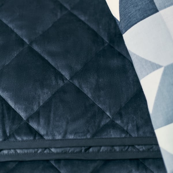 Un couvre-lit matelassé MJUKPLISTER gris foncé derrière un coussin avec une housse SVARTHÖ bleu / vert.