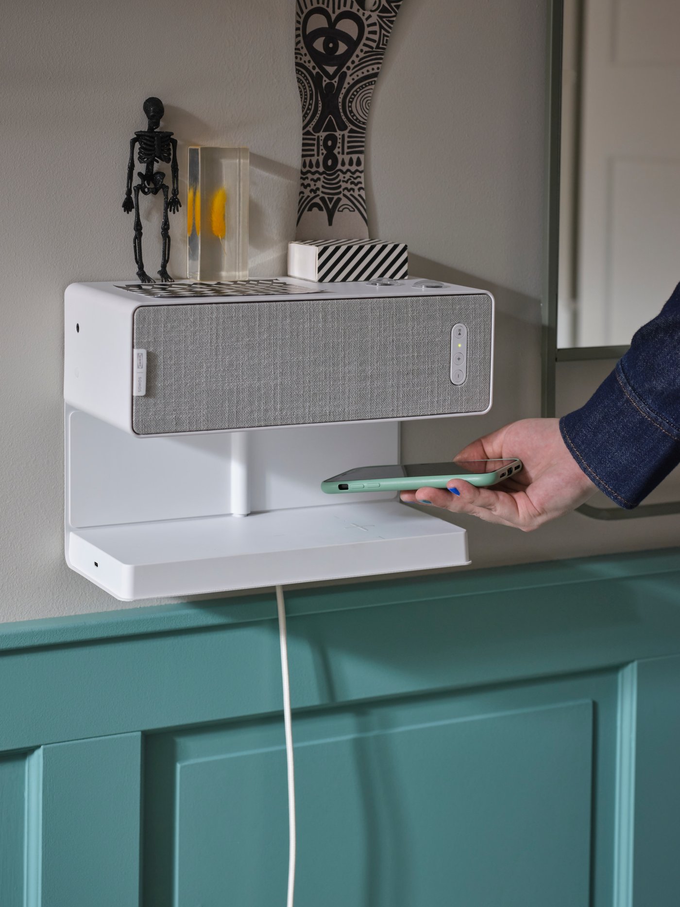 A white SYMFONISK shelf with wireless charger and SYMFONISK bookshelf speaker on a wall. A person holds a smartphone by them.