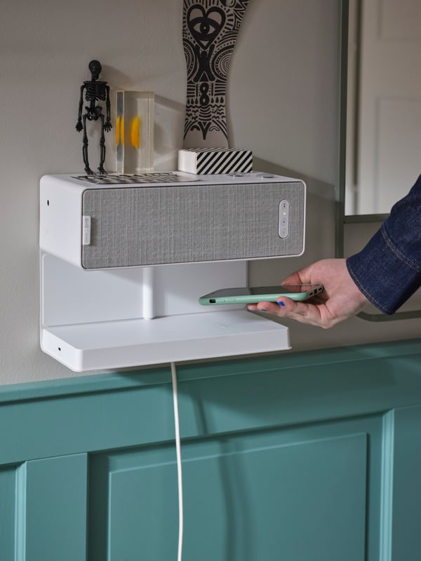Une main en train de déposer un téléphone portable sur une tablette avec chargeur sans fil; une enceinte étagère WiFi SYMFONISK blanche est montée sur la tablette.