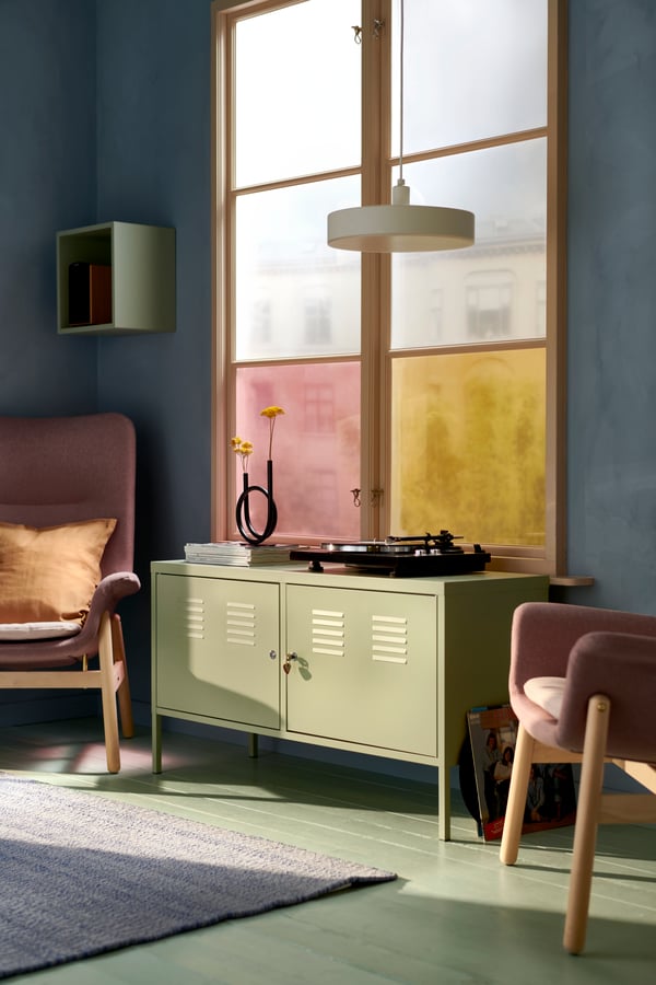 Ett IKEA PS skåp står mitt emellan två rosa VEDBO fåtöljer i ett rum med ett stort fönster och blåa väggar.