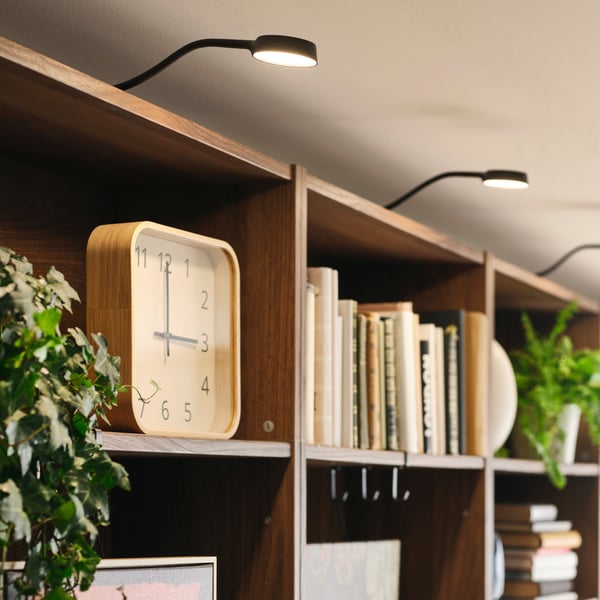 Reloj de pared SKOGSSVALA con acabado en chapa de roble, colocado sobre una repisa con libros y plantas.