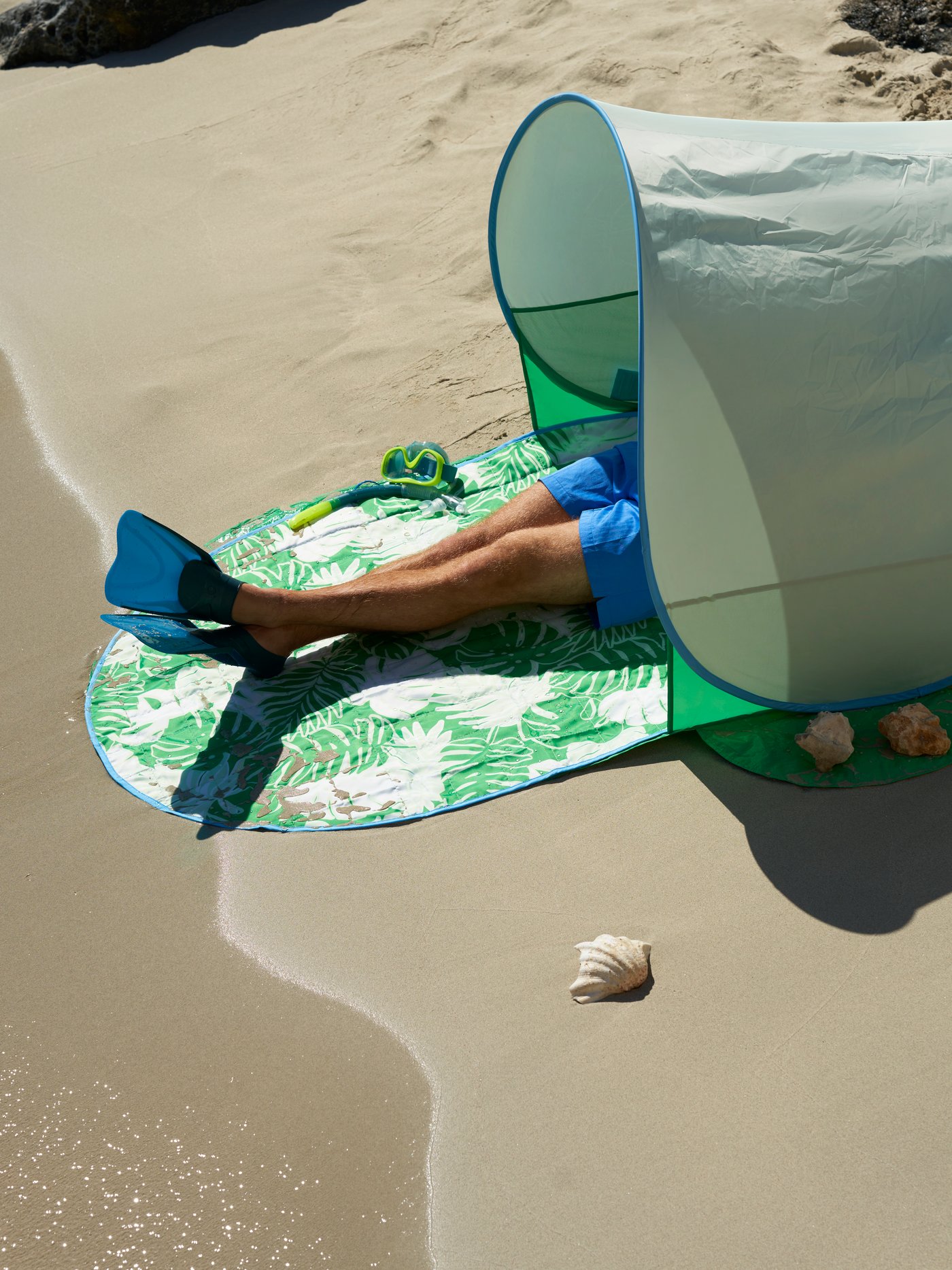 En person ligger på et tæppe på stranden med et skyggetelt over sig. 