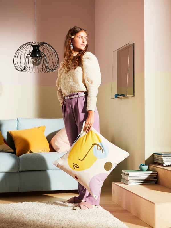 A woman holding a MANDELPIL cushion with an abstract pattern, standing in a pastel living room with a unique pendant lampshade.