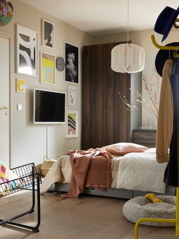 A grey GLADSTAD bed with green and cream textiles on it, in front of a PAX/STORKLINTA wardobe and opposite a picture wall.