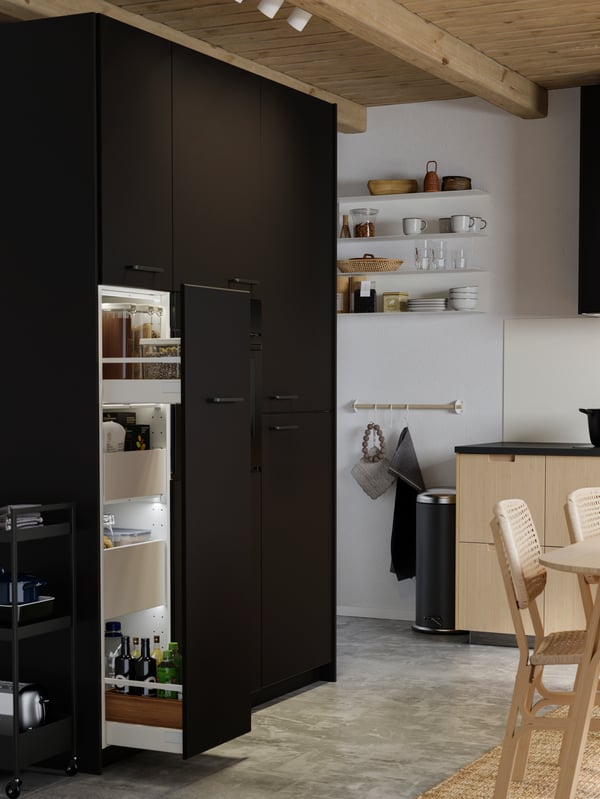 An anthracite pull-out pantry with bottles of oil and transparent containers with pasta, and two light bamboo chairs.