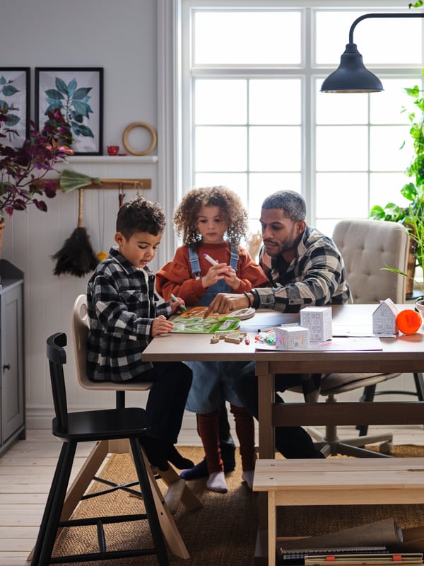 A man and two young children, pens in hand, absorbed in a playful project around a grey-brown KLIMPFJÄLL dining table.