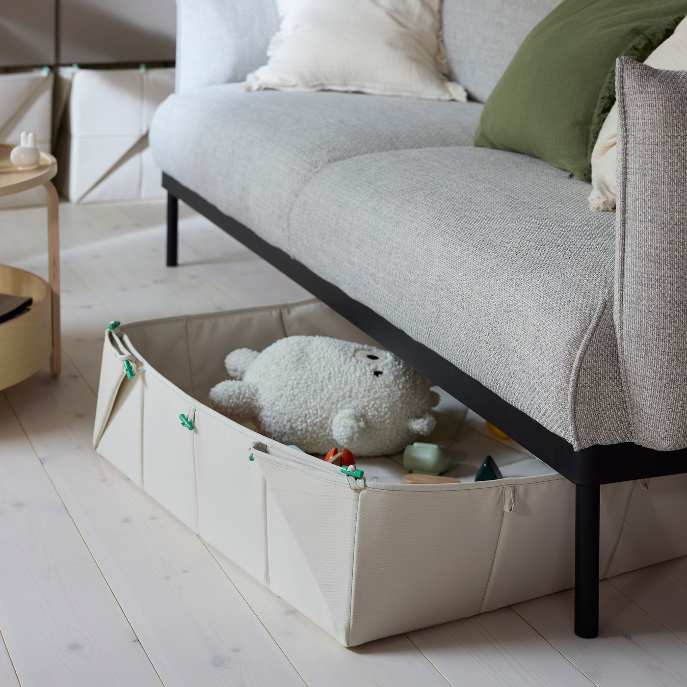 A beige GREJSIMOJS mat/foldable storage box pokes out from under a sofa in a modern living room. Soft toys are in the box.