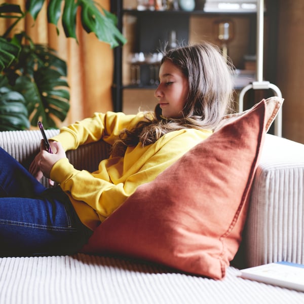 A girl looking at her phone while comfortably reclining on a grey/beige JÄTTEBO 4.5-seat modular sofa with chaise longue.