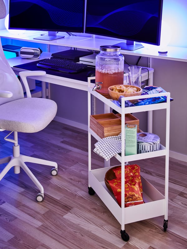 Un carrello con rotelle bianco su cui ci sono snack e una bevanda accanto a una sedia da ufficio e ripiani con due schermi per computer.