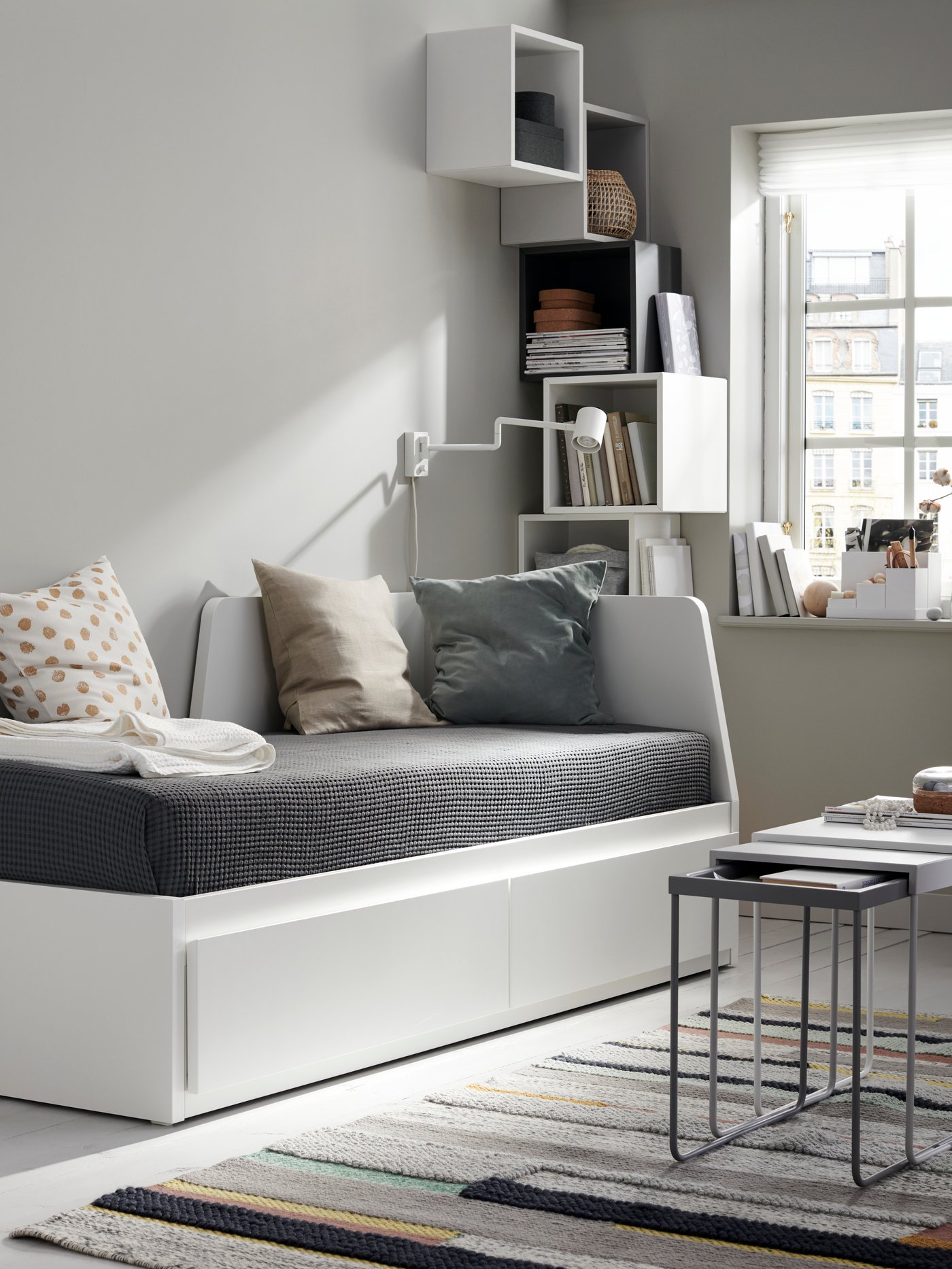A white FLEKKE day-bed with two drawers and two mattresses in a room with a nest of tables, a handwoven rug and a wall lamp.