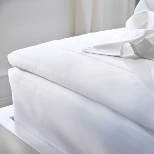 Close-up of corner of a bedframe, mattress and mattress pad with sheets in a pile on top, all in crisp white.