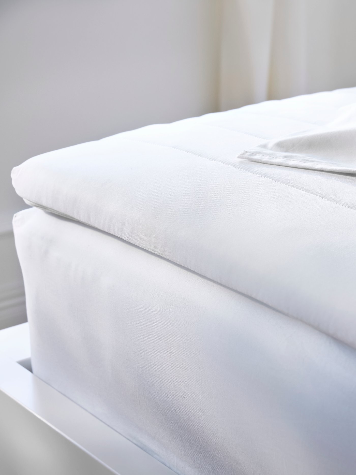 Gros plan sur le coin d’une structure de lit avec matelas et surmatelas sur lequel est posée une pile de draps, le tout d’un blanc éclatant.