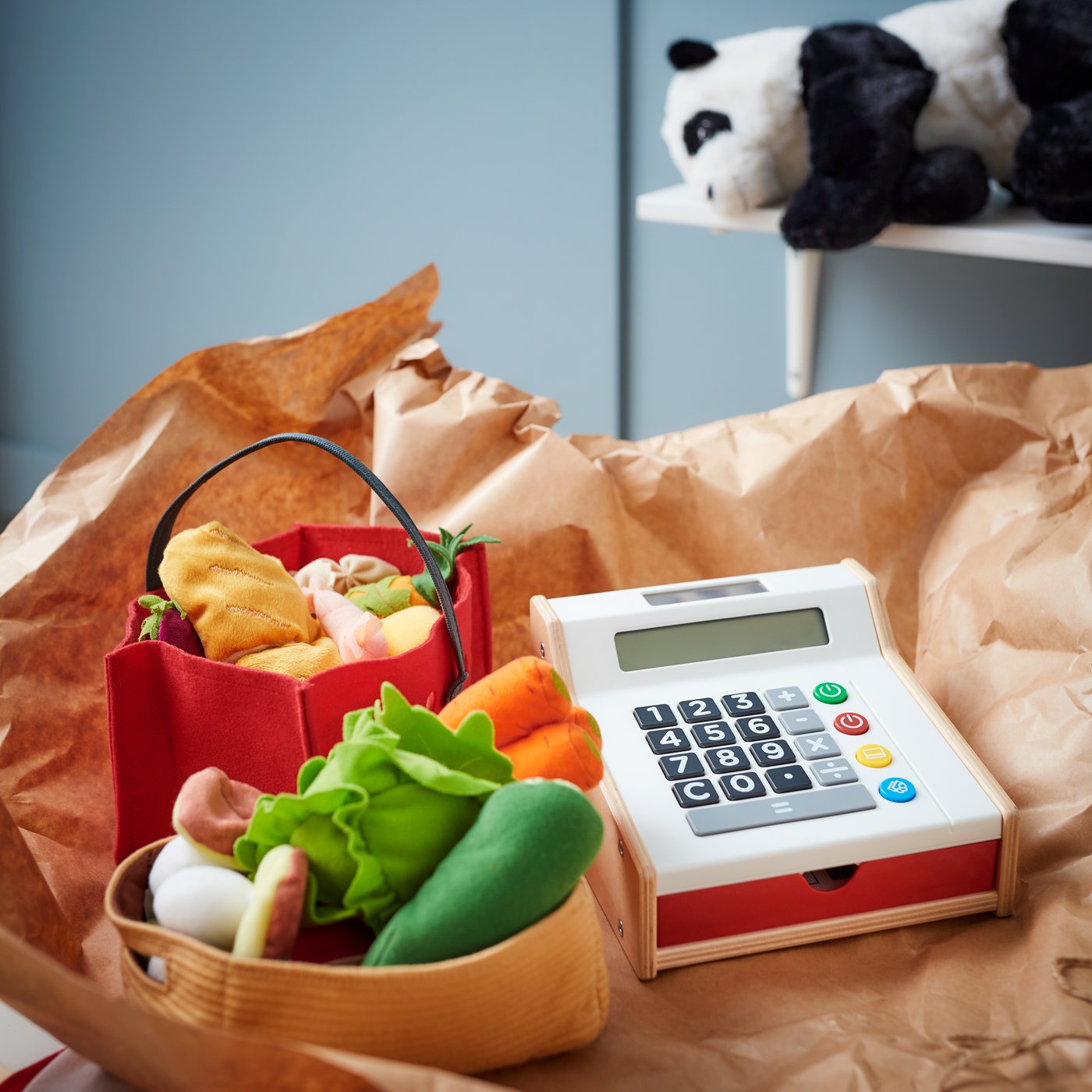 Caisse enregistreuse jouet DUKTIG et quelques légumes en peluche DUKTIG posés sur une feuille de papier kraft.