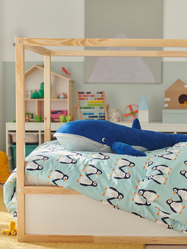 A BLÅVINGAD soft toy blue whale lies on a duvet with a BLÅVINGAD duvet cover on a KURA reversible bed.