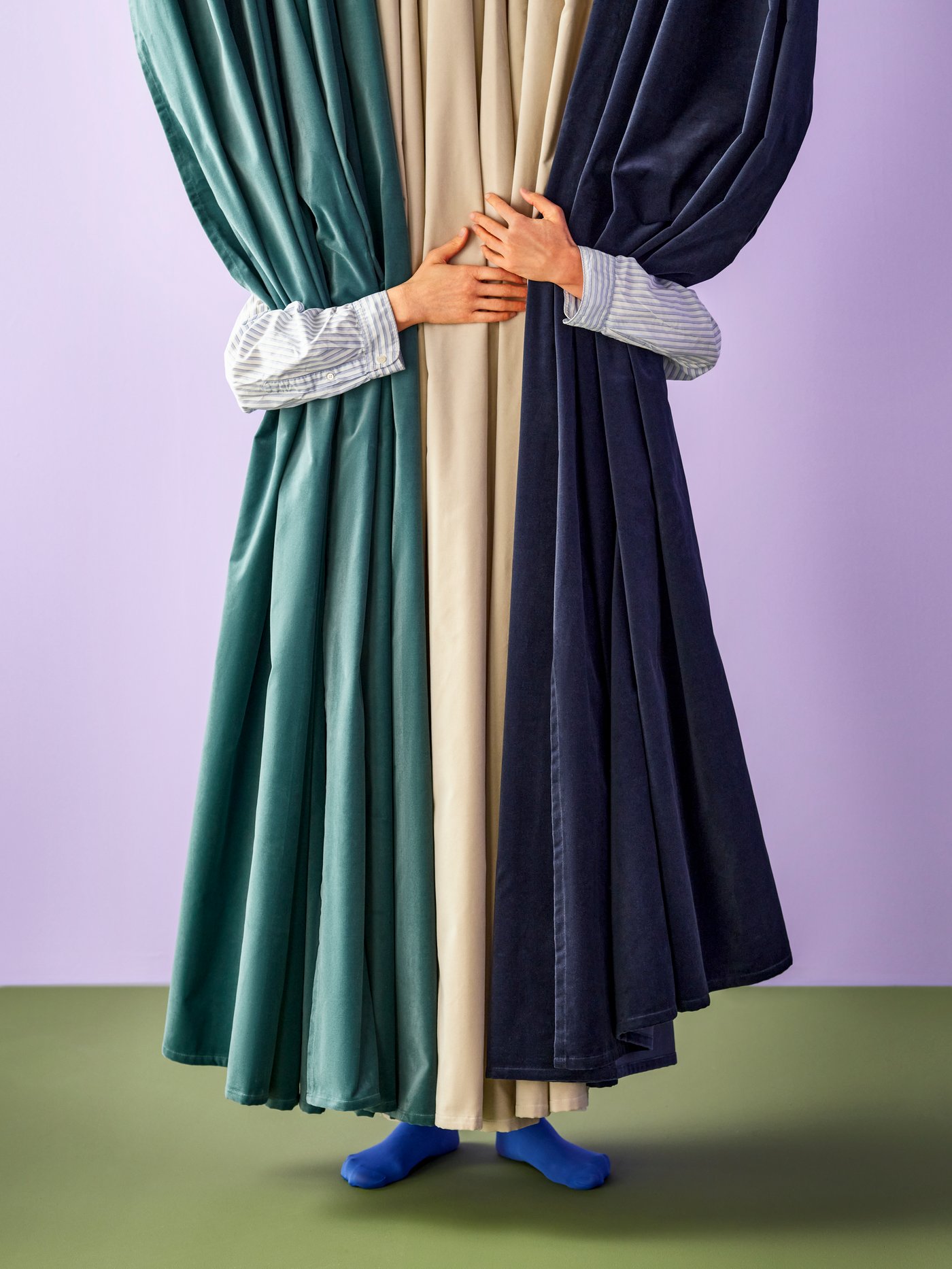 A person embracing three SANELA curtains from behind. The curtains are dark blue, dark grey-turquoise and light beige.