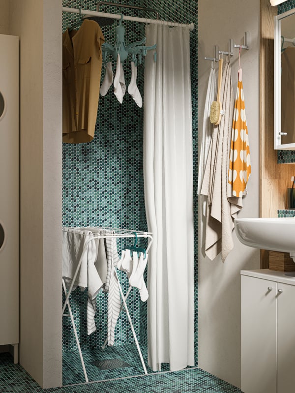 A shower area with green tiles, a drying rack with socks and shirts hanging, a white shower curtain, a shirt on a hanger.