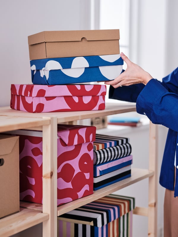 A person places two boxes, one covered with RAKLOSTA pre-cut fabric, on a box covered with pink FLUGBLOMSTER pre-cut fabric.