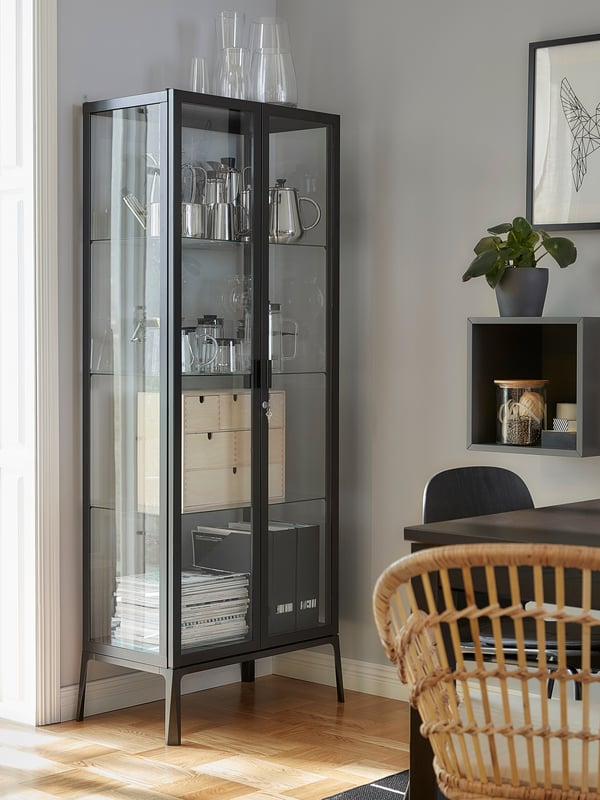 A MILSBO glass-door cabinet stands next to a balcony door. Decorative items, magazines and magazine files are stored inside.