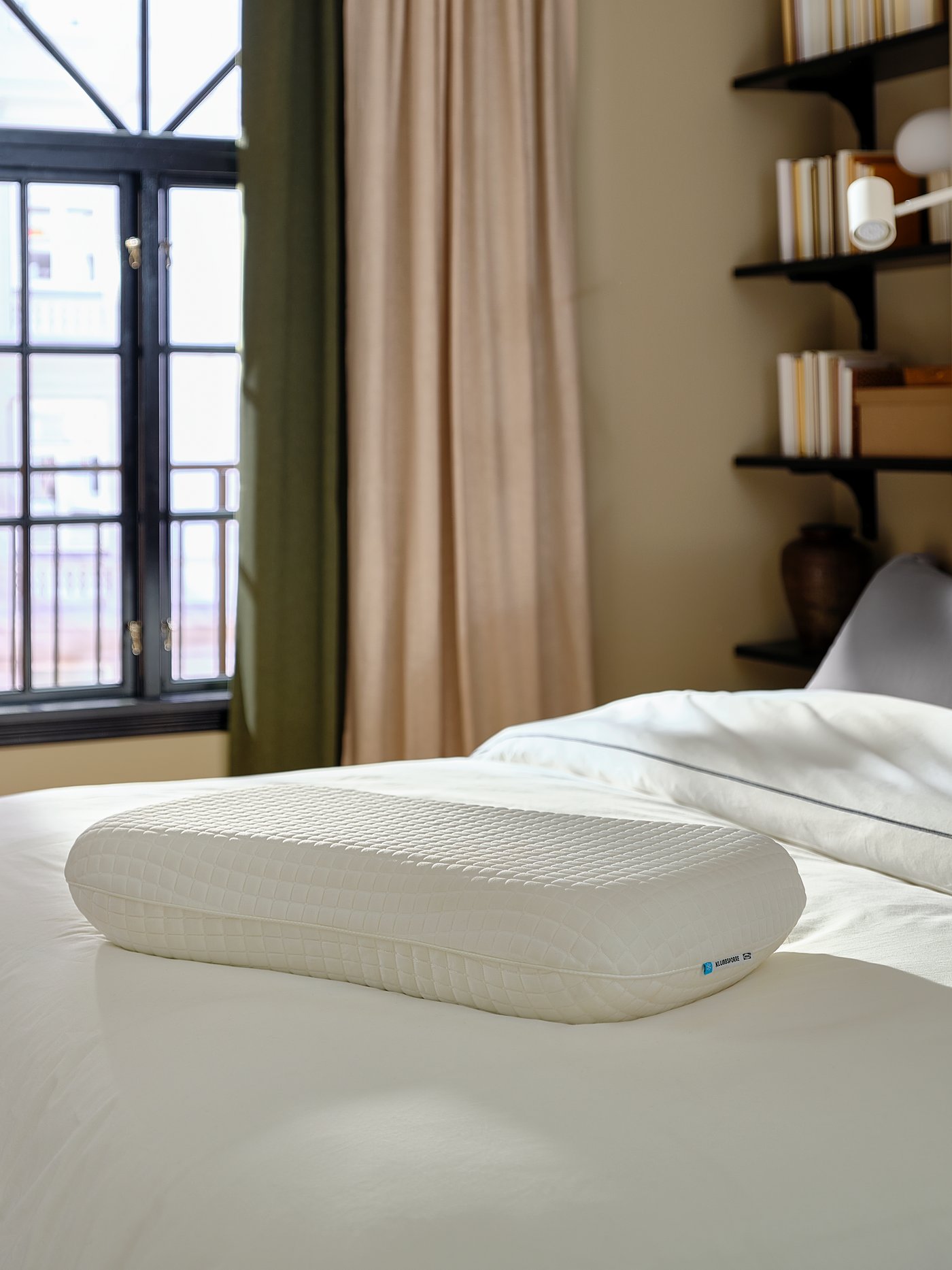 Ergonomic pillow on a white bed beside a window.