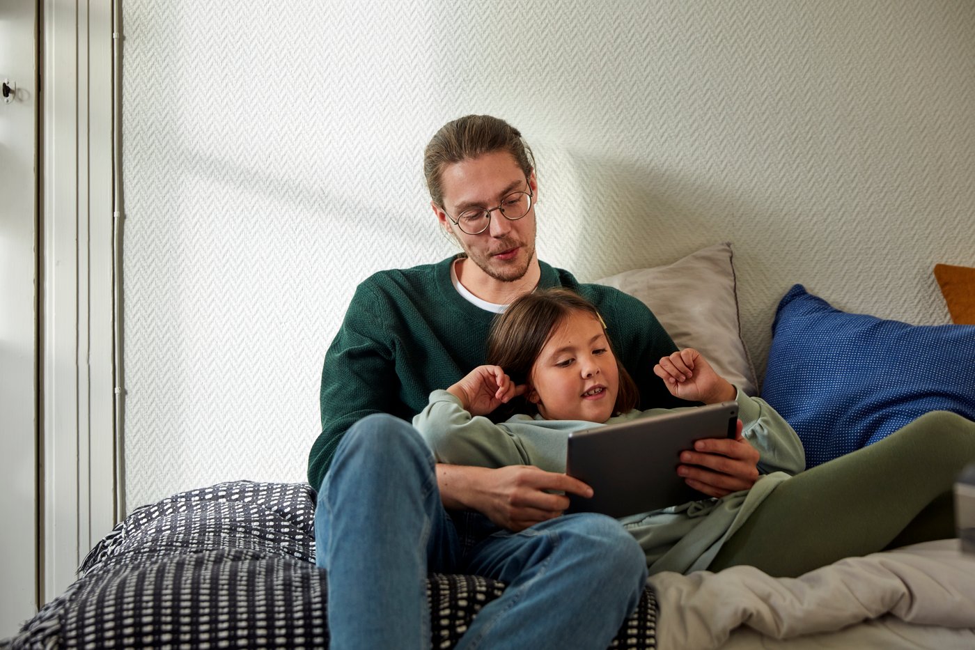 Une personne assise sur un lit avec un enfant regardant une tablette.