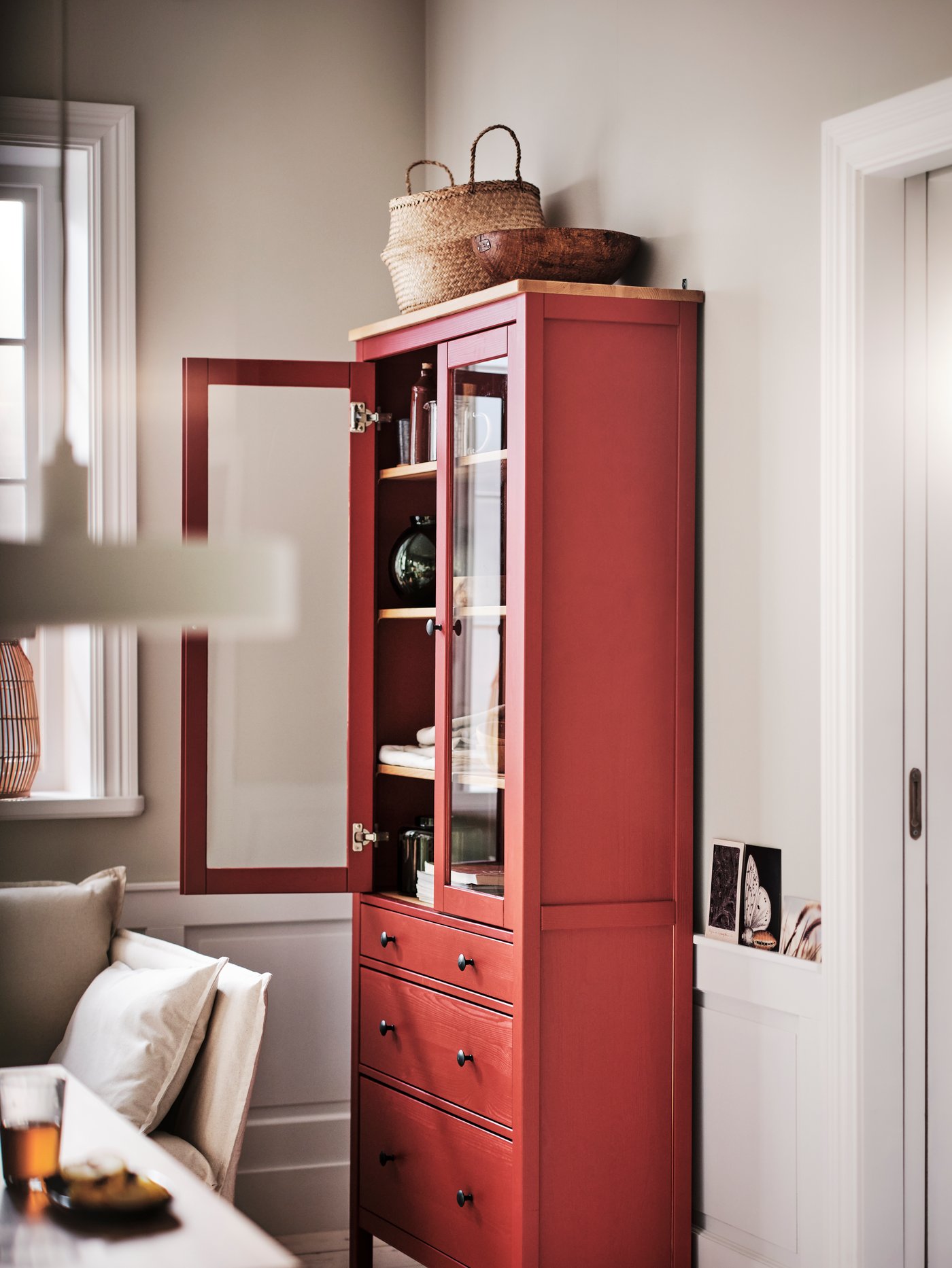 A red stain/light brown HEMNES glass-door cabinet with 3 drawers with one door open and a seagrass FLÅDIS basket on top.