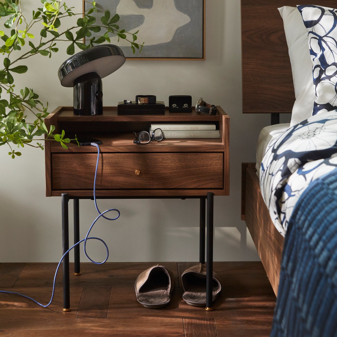 Une chambre avec table de chevet RÅDMANSÖ brune avec pieds effet noyer, à côté d’un lit et d’une plante en pot.