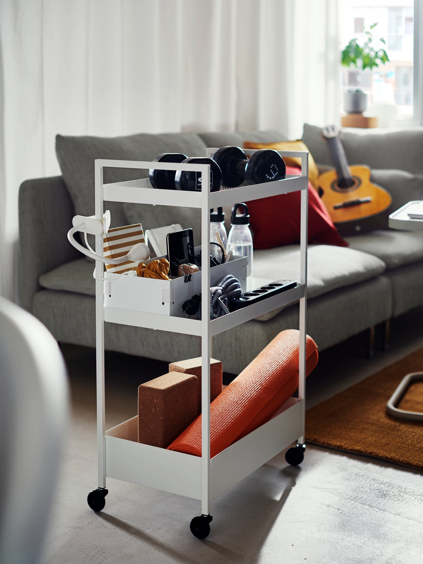 Bílý pojízdný vozík IKEA NISSAFORS v obývacím pokoji, naplněný fitness vybavením: činky, láhve na vodu, jóga podložka, cvičební bloky a drobné doplňky. V pozadí šedá pohovka s polštáři, kytara a konferenční stolek.