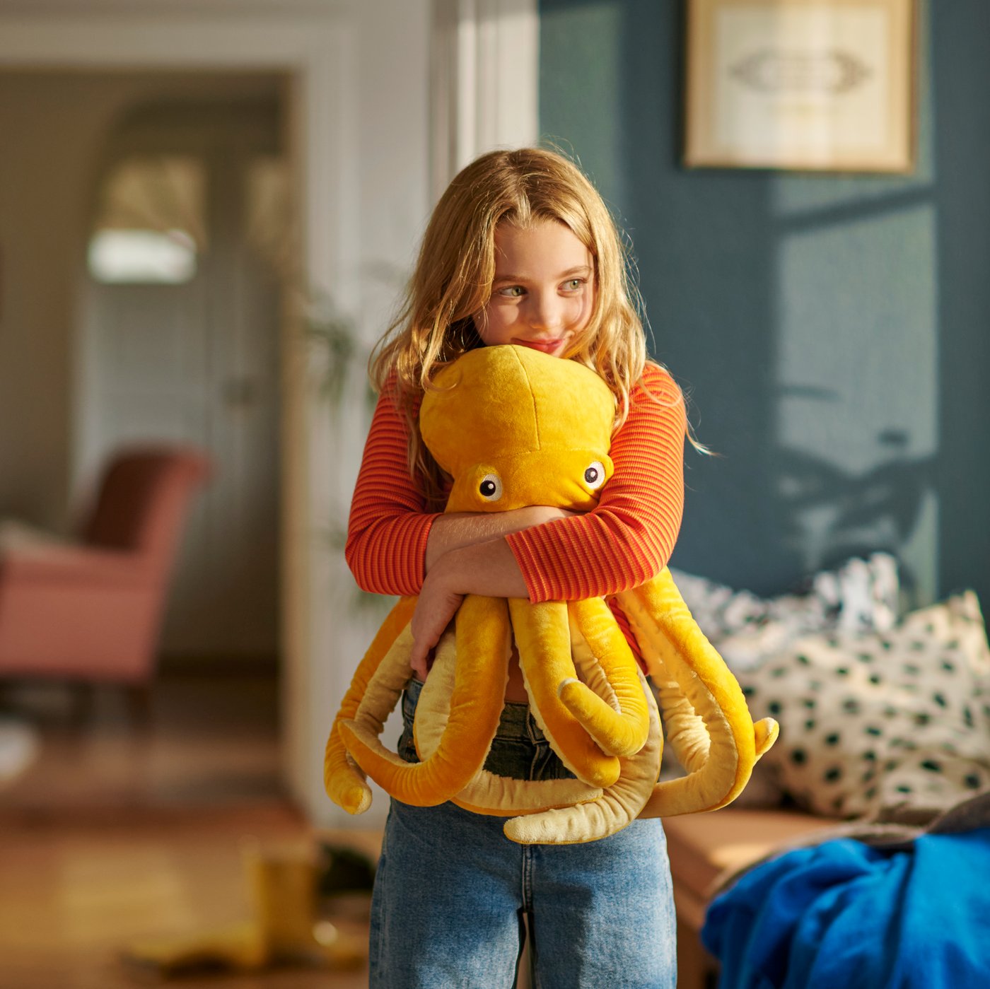 A child is standing at home squeezing tightly a yellow BLÅVINGAD octopus soft toy.