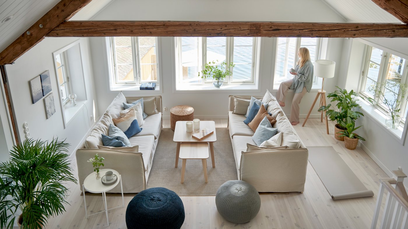 Ljust och luftigt vardagsrum med stora fönster, två beige BACKSÄLEN soffor i L-form runt ett ljust träsoffbord, blå och beige kuddar, gröna växter, två sittpuffar i blått och grått, och en person som sitter i fönstret med en kaffe i handen.