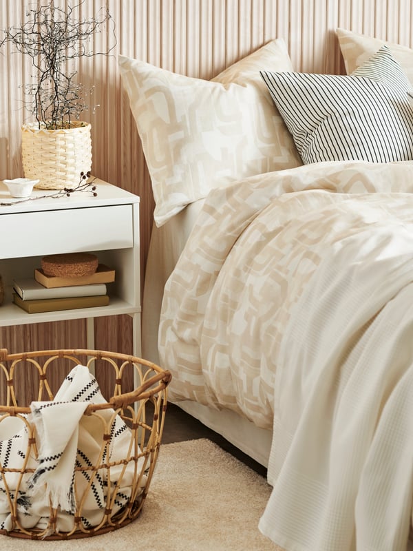 An ESPEVÄR white divan bed with patterned bed linen and a striped throw pillow. A white bed side table sits next to the bed with a rattan basket on a rug.