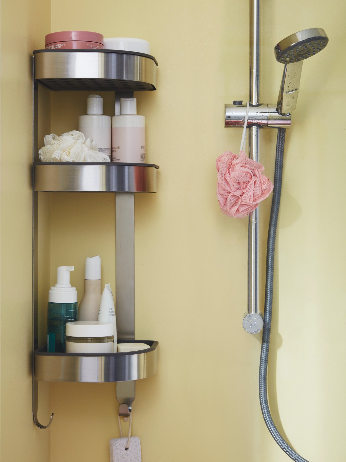 A metal IKEA shower shelf hangs on a soft yellow wall, holding shampoo bottles, soap, a sponge, and personal-care products beside a handheld shower with a pink bath puff.
