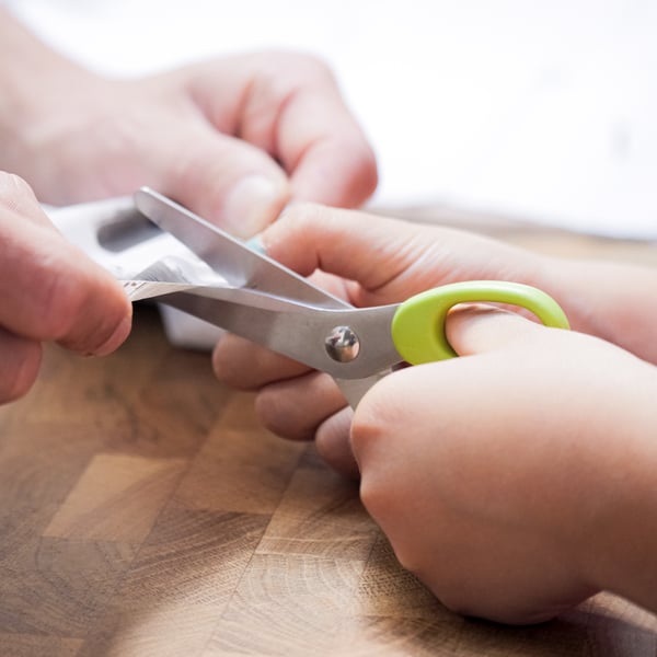 Tijeras MÅLA con bordes verdes y usadas por una mano infantil.