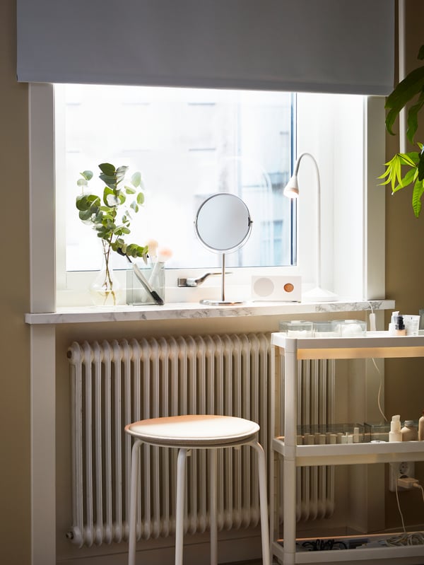 Una habitación iluminada de tonos crema donde se ve un carrito organizador y un piso. En la ventana se ve una cortina roller gris a medio bajar.