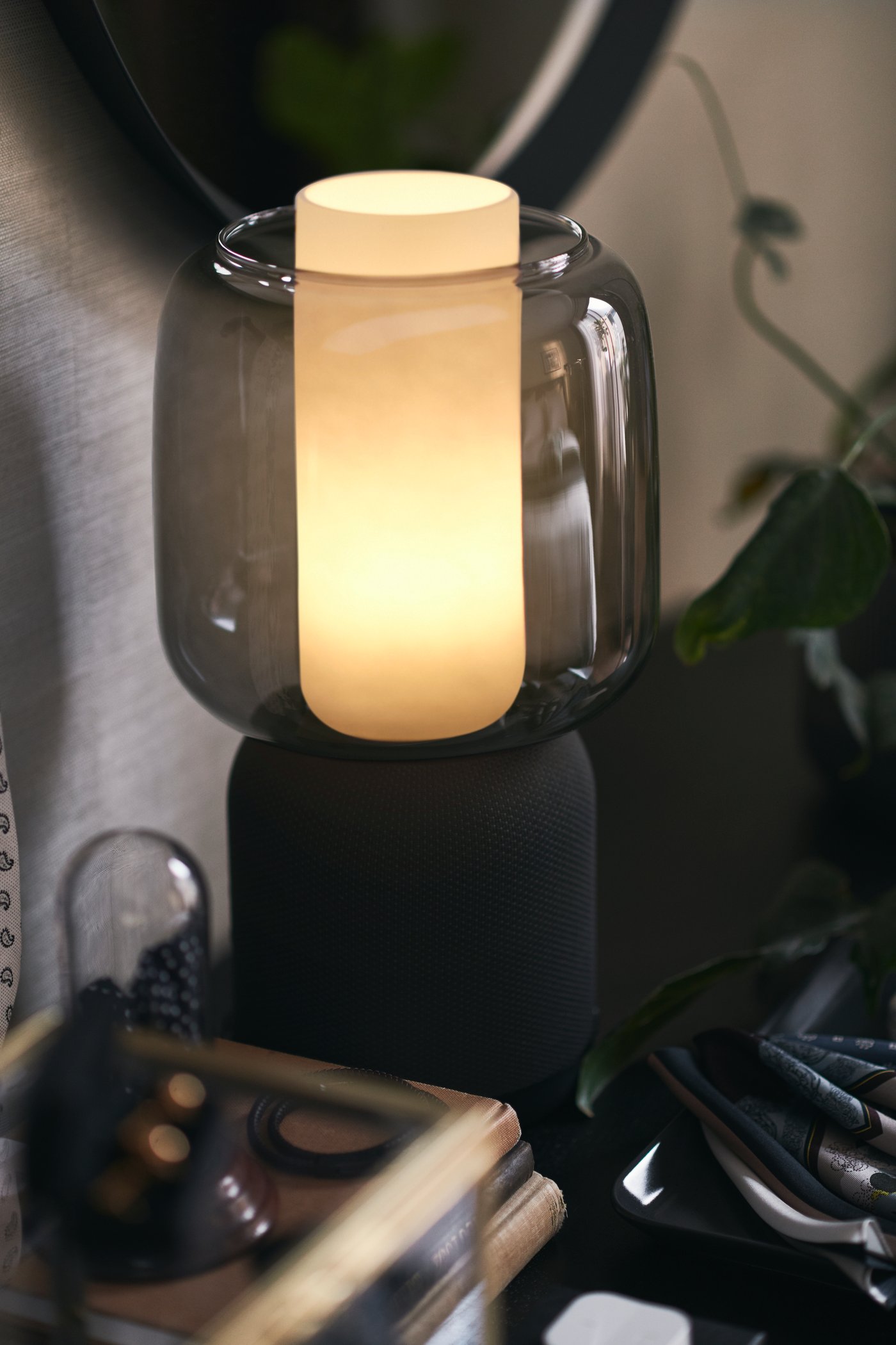 Accanto a una parete grigia, una lampada/cassa Wi-Fi SYMFONISK nera con un paralume in vetro, con dietro una pianta rampicante - IKEA