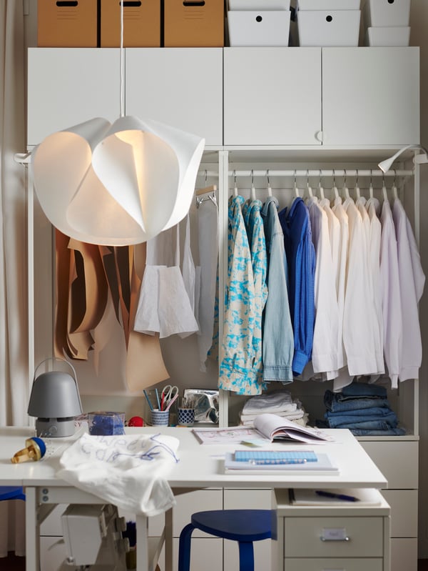 Un bureau est installé devant une combinaison armoire-penderie PLATSA dans laquelle sont rangés des vêtements sur cintres, dont des articles en cours de réalisation.