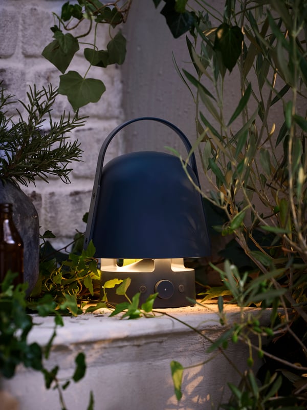 A patio at dusk with a blue VAPPEBY outdoor bluetooth speaker lamp placed in a corner. It spreads a soft and cosy light.