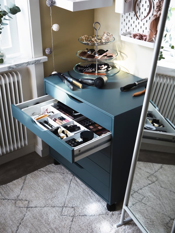 Un caisson à tiroirs ALEX gris turquoise avec un tiroir ouvert et des articles pour cheveux à l’intérieur et sur le caisson, et un miroir sur pied blanc.