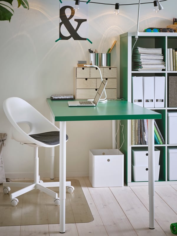 Study space with a green table with white legs, a swivel chair, a white table lamp and a transparent floor protector.
